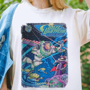 Puede incluir: Camiseta blanca con un gr&aacute;fico de estilo vintage de Buzz Lightyear de la franquicia Toy Story. El gr&aacute;fico presenta a Buzz en su traje de ranger espacial, volando por el espacio con un disparo l&aacute;ser. El texto "Buzz Lightyear Laser Blast" est&aacute; impreso sobre el gr&aacute;fico, y "Discoveryland" est&aacute; impreso debajo.