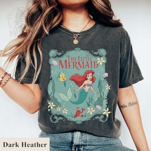 May include: Dark heather gray t-shirt featuring a graphic of Ariel from The Little Mermaid. The design includes Ariel, Flounder, Sebastian, and the words "The Little Mermaid" in a decorative frame. The shirt is a vintage style.