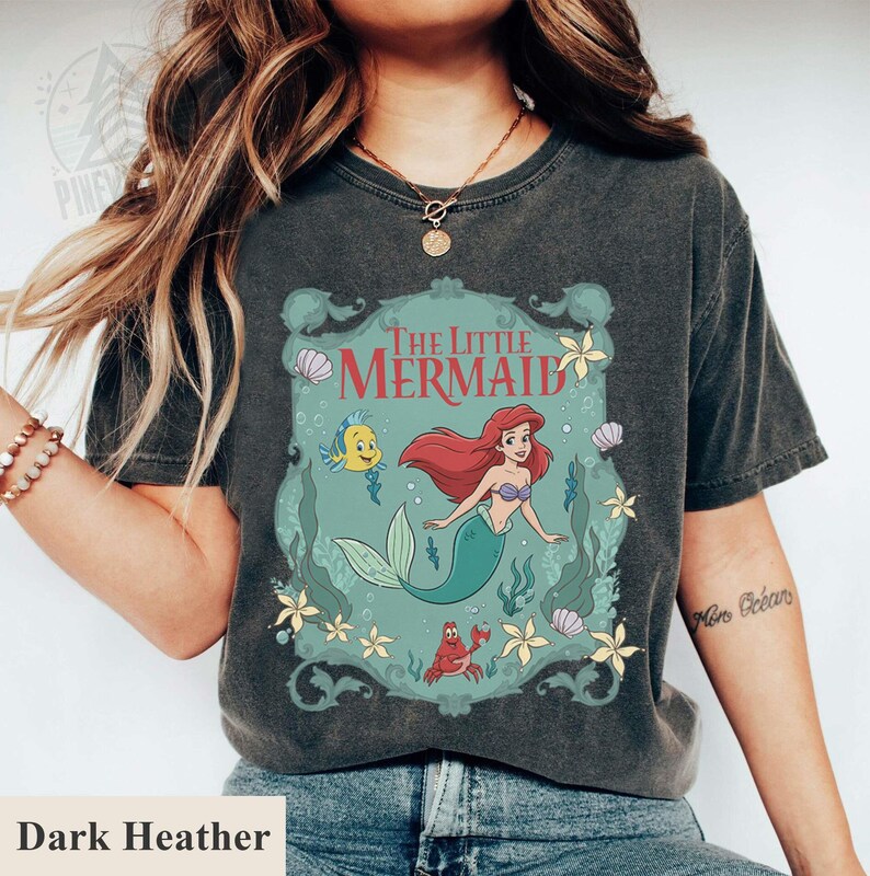 May include: Dark heather gray t-shirt featuring a graphic of Ariel from The Little Mermaid. The design includes Ariel, Flounder, Sebastian, and the words "The Little Mermaid" in a decorative frame. The shirt is a vintage style.