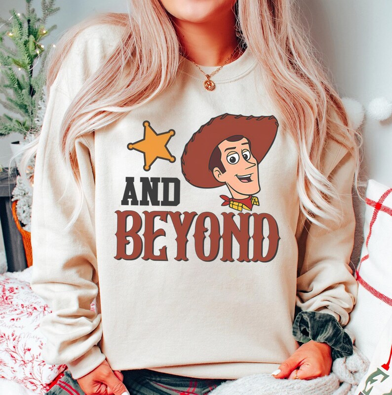 May include: A beige sweatshirt featuring a cartoon image of a sheriff star and a cowboy character with the text "AND BEYOND" in brown letters. The cowboy has a brown hat, a red neckerchief, and a yellow shirt.