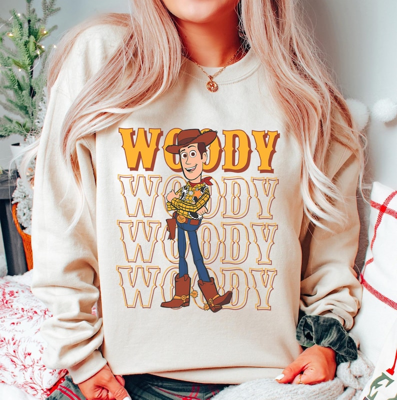 Puede incluir: Una sudadera beige con un gr&aacute;fico del personaje Woody de Toy Story. La palabra "Woody" est&aacute; impresa en letras grandes de color naranja encima del personaje, con el nombre repetido debajo. Woody est&aacute; representado con su atuendo de vaquero caracter&iacute;stico.