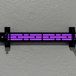 Puede incluir: Una l&aacute;mpara de pared negra con una serie de luces p&uacute;rpuras horizontales. La l&aacute;mpara es rectangular con una serie de recortes. La l&aacute;mpara est&aacute; montada en una pared gris texturizada.