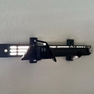 Puede incluir: Soporte de exhibici&oacute;n de sable de luz negro con un sable de luz a juego. El soporte est&aacute; fijado a una pared y presenta una serie de luces rectangulares. El sable de luz es negro con detalles plateados.