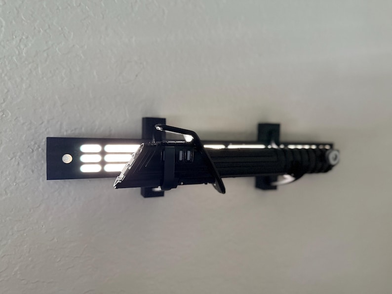 Puede incluir: Soporte de exhibici&oacute;n de sable de luz negro con un sable de luz a juego. El soporte est&aacute; fijado a una pared y presenta una serie de luces rectangulares. El sable de luz es negro con detalles plateados.
