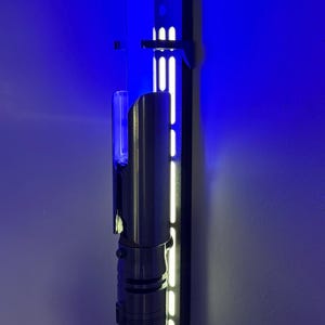 May include: A silver and black lightsaber is mounted on a wall with a black holder. The wall is painted a deep blue, and the lightsaber is illuminated with a blue glow. The holder has vertical white lights.