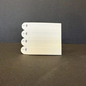 Peut inclure: Une pile de quatre bandes adhésives rectangulaires blanc cassé avec des bords arrondis. Chaque bande porte une petite flèche et le logo "3M". Les bandes sont disposées verticalement sur un fond sombre.