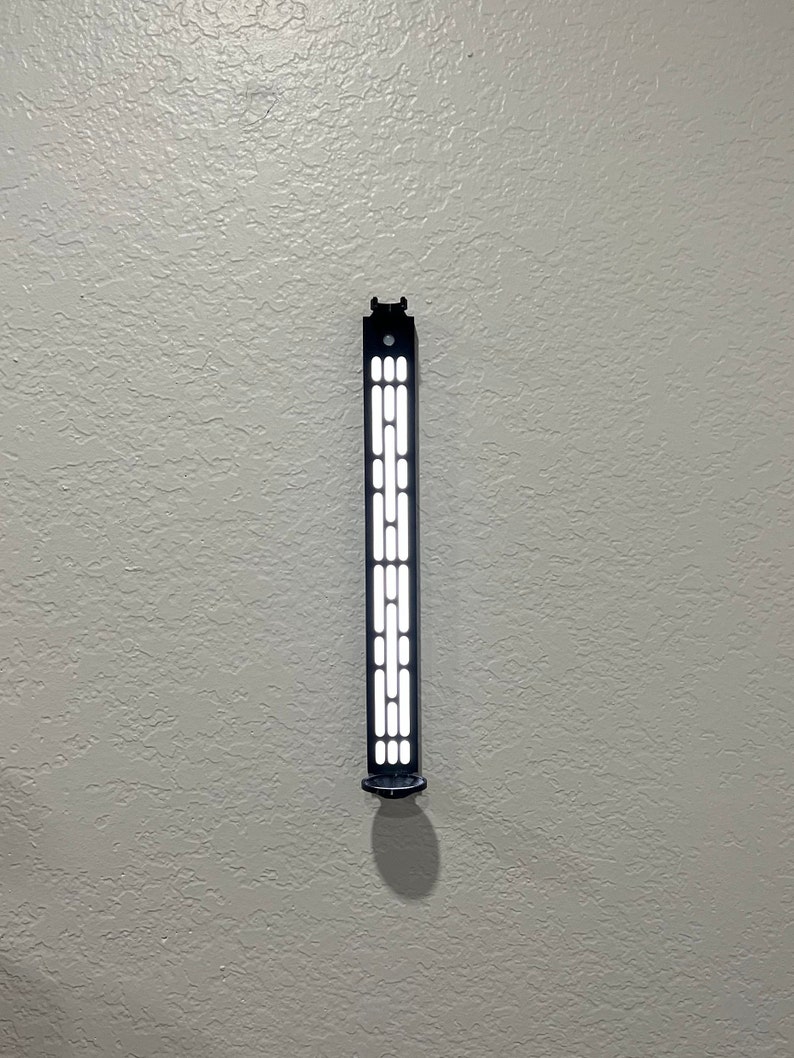 May include: A black wall-mounted light fixture with a rectangular design. The fixture has a series of rectangular and circular cutouts that emit light. The fixture is mounted on a textured, light gray wall.