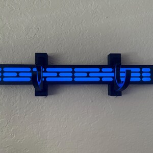 Puede incluir: Un perchero negro montado en la pared con dos ganchos curvos. El perchero presenta una serie de formas rectangulares y ovaladas azules iluminadas. El dise&ntilde;o est&aacute; montado en una pared de color claro.