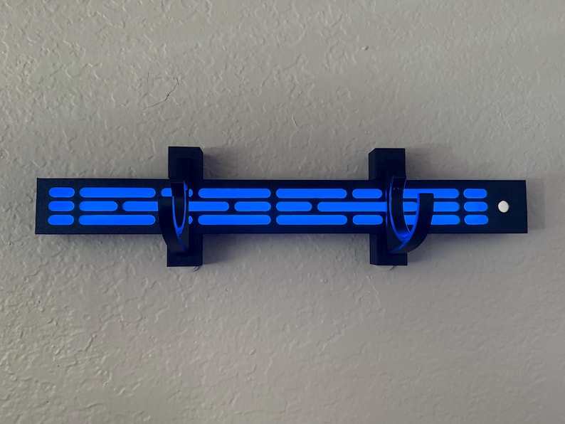 Puede incluir: Un perchero negro montado en la pared con dos ganchos curvos. El perchero presenta una serie de formas rectangulares y ovaladas azules iluminadas. El dise&ntilde;o est&aacute; montado en una pared de color claro.