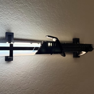 Puede incluir: Un sable de luz negro montado en una pared. El sable de luz tiene una hoja blanca con una franja negra y una empu&ntilde;adura negra. El sable de luz se sujeta con dos soportes de pared negros.