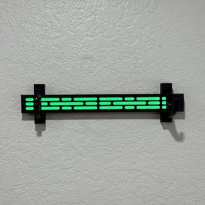 Puede incluir: Una l&aacute;mpara de pared negra con un brillo verde ne&oacute;n. La l&aacute;mpara tiene una serie de recortes rectangulares horizontales que emiten luz. La l&aacute;mpara est&aacute; montada en una pared texturizada de color blanco roto.
