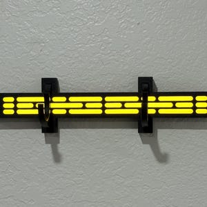 Puede incluir: Un perchero de pared negro con un dise&ntilde;o brillante amarillo. El perchero tiene dos ganchos negros y una serie de rect&aacute;ngulos y puntos amarillos horizontales. El dise&ntilde;o recuerda un tema de ciencia ficci&oacute;n.