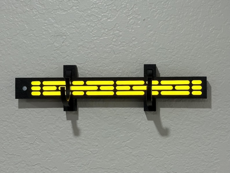 Puede incluir: Un perchero de pared negro con un dise&ntilde;o brillante amarillo. El perchero tiene dos ganchos negros y una serie de rect&aacute;ngulos y puntos amarillos horizontales. El dise&ntilde;o recuerda un tema de ciencia ficci&oacute;n.