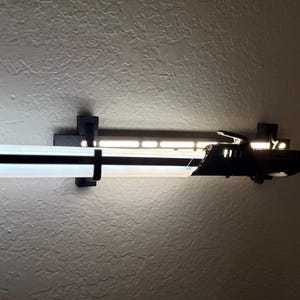 Puede incluir: Una moderna l&aacute;mpara LED en blanco y negro montada en una pared texturizada. La l&aacute;mpara tiene forma rectangular con una tira de luz blanca brillante y un acento negro. Se sujeta con soportes negros.