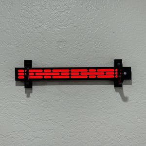 Puede incluir: Una l&aacute;mpara de pared negra con una serie de luces rojas rectangulares horizontales. La l&aacute;mpara est&aacute; montada en una pared gris claro texturizada. La l&aacute;mpara mide aproximadamente 30 cm de largo.
