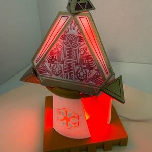 Könnte beinhalten: Eine geometrische, beleuchtete Lampe mit rotem Leuchten. Die Lampe hat eine pyramidenförmige Oberseite mit komplizierten Designs und eine kleinere Pyramide oben. Die Basis hat eine rechteckige Form mit einem weißen Zylinder und einem goldenen Rand.