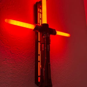 May include: A wall-mounted lightsaber lamp in black and red. The lamp has a vertical central light and a horizontal blade, both glowing red. The design includes a detailed hilt and mounting bracket, creating a striking visual effect.