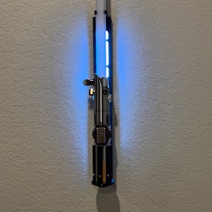 May include: A silver and black lightsaber with a blue glowing blade is mounted on a textured wall. The hilt features metallic details and a red button. The blade is illuminated, creating a striking visual effect.