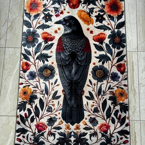 May include: Rectangular rug with a detailed illustration of a black crow with red accents, framed by a floral border. The flowers are in shades of orange, red, and blue against a cream background. The rug is placed on a tiled floor.
