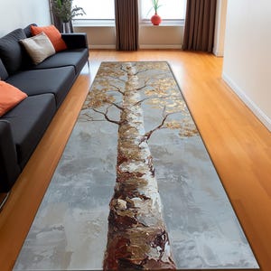 May include: A rectangular area rug featuring a painted tree design. The tree trunk is brown and white, with gold-colored leaves and branches against a gray background. The rug is designed for indoor use.