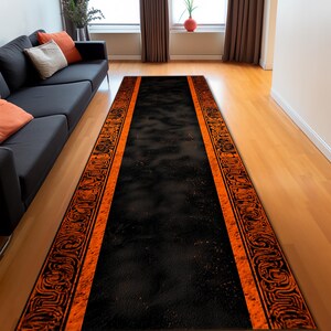 Könnte beinhalten: Ein langer, rechteckiger schwarzer Teppich mit orangefarbenen Rändern und dekorativen Mustern. Der Teppich liegt auf einem Holzboden in einem Wohnzimmer, mit einem schwarzen Sofa und Fenstern im Hintergrund.