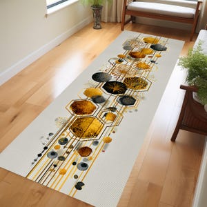 May include: A rectangular, off-white runner rug with a geometric design. The pattern features gold, black, and brown hexagonal shapes and circular accents connected by gold lines. The rug is placed on a hardwood floor.