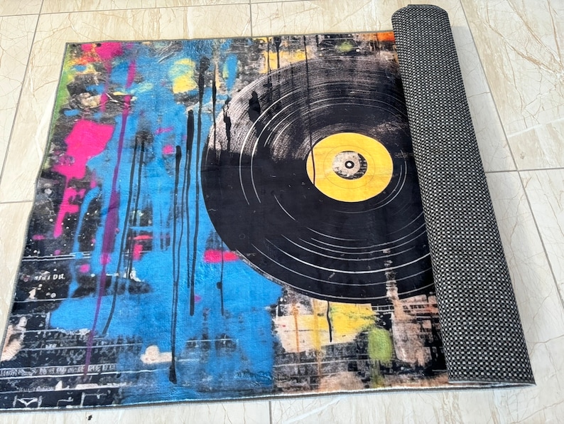 K&ouml;nnte beinhalten: Ein bunter Teppich mit einem gro&szlig;en schwarzen und gelben Vinyl-Schallplatten-Design. Der Teppich hat ein abstraktes Spritzmuster in Blau, Pink und Gelb. Der Teppich ist aufgerollt und hat eine schwarze R&uuml;ckseite.
