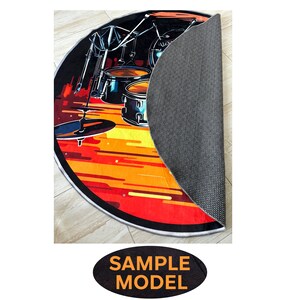May include: Round rug with a drum set illustration. The design features a drum kit in black and blue against a backdrop of orange and yellow stripes. The rug's edge is black with a white border. The rug is partially rolled up, revealing a gray, textured underside.