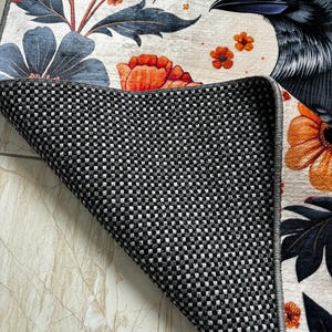 May include: A white rug with a black raven, orange and blue flowers, and green leaves. The rug has a black and white checkered backing.