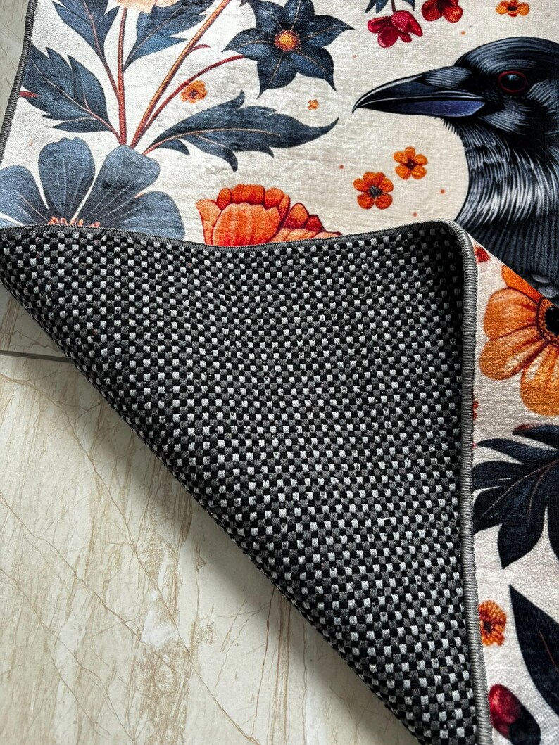 May include: A white rug with a black raven, orange and blue flowers, and green leaves. The rug has a black and white checkered backing.