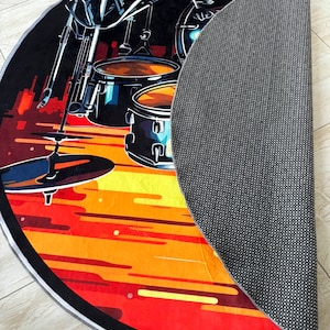May include: A round area rug featuring a drum set illustration. The rug has a black background with a vibrant orange and yellow sunset design. The drum set is depicted in shades of blue and black.