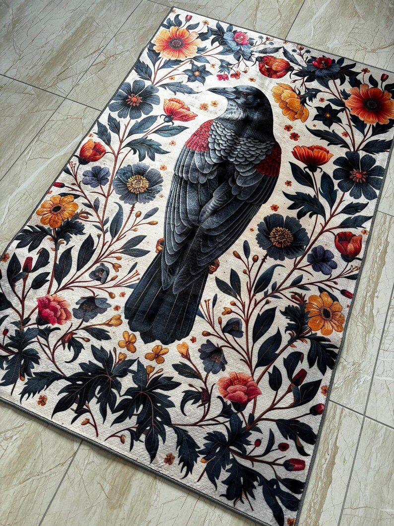 May include: Rectangular rug with a detailed illustration of a black bird with red accents, surrounded by a floral pattern of orange, blue, and pink flowers on a white background. The rug has a grey border.