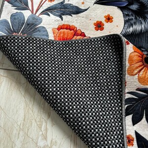 May include: A white rug featuring a black raven, orange and blue flowers, and green leaves. The rug has a black and white checkered backing.