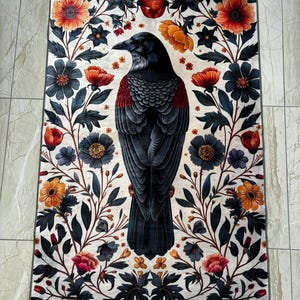 May include: A black raven with a red breast is depicted in the center of a white rug with a floral pattern. The flowers are in shades of red, orange, yellow, and blue.
