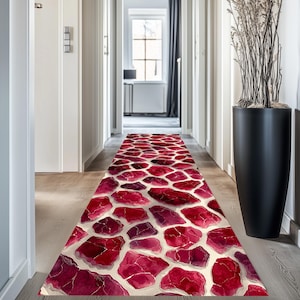 Może przedstawiać: Dywanik do przedpokoju z czerwono-białym wzorem kamieni. Dywanik ma nieregularne czerwone kształty ze złotymi akcentami na białym tle. Dywanik jest umieszczony na jasnej drewnianej podłodze, dodając dekoracyjny akcent do przedpokoju.
