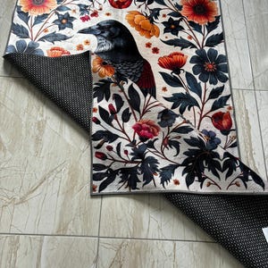 May include: A white rug with a black raven, red and orange flowers, and green leaves. The rug has a black non-slip backing.