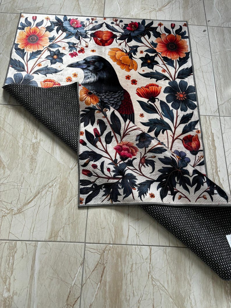 May include: A white rug with a black raven, red and orange flowers, and green leaves. The rug has a black non-slip backing.