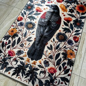 May include: A black raven with red accents stands in the center of a white rug with a floral pattern. The flowers are in shades of red, orange, yellow, and blue. The rug has a gray border.