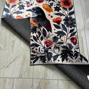 May include: A rectangular rug with a detailed illustration of a crow amidst floral patterns in orange, red, blue, and black. The design is set against a cream background. The underside of the rug is black with white dots.