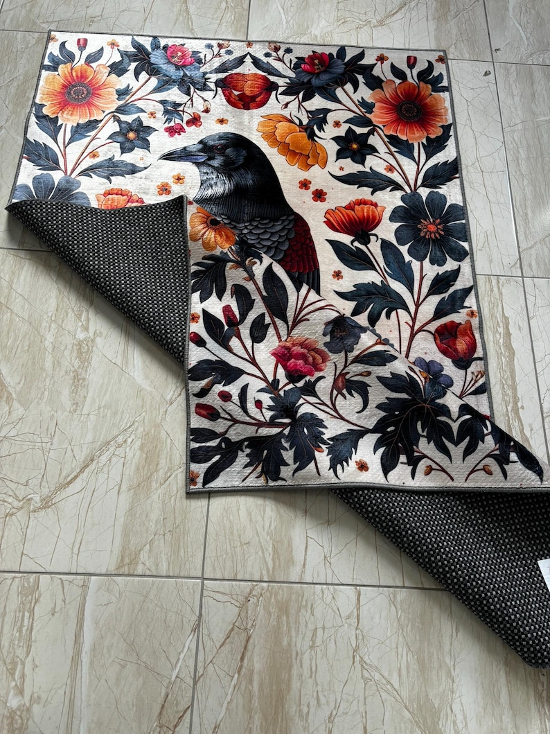 May include: A rectangular rug with a detailed illustration of a crow amidst floral patterns in orange, red, blue, and black. The design is set against a cream background. The underside of the rug is black with white dots.