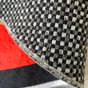 May include: Close-up of a section of a rug with a geometric pattern. The top portion features a black, white, and gray checkered design. The bottom section has a red, black, and white striped pattern. The edges are finished with a stitched border.