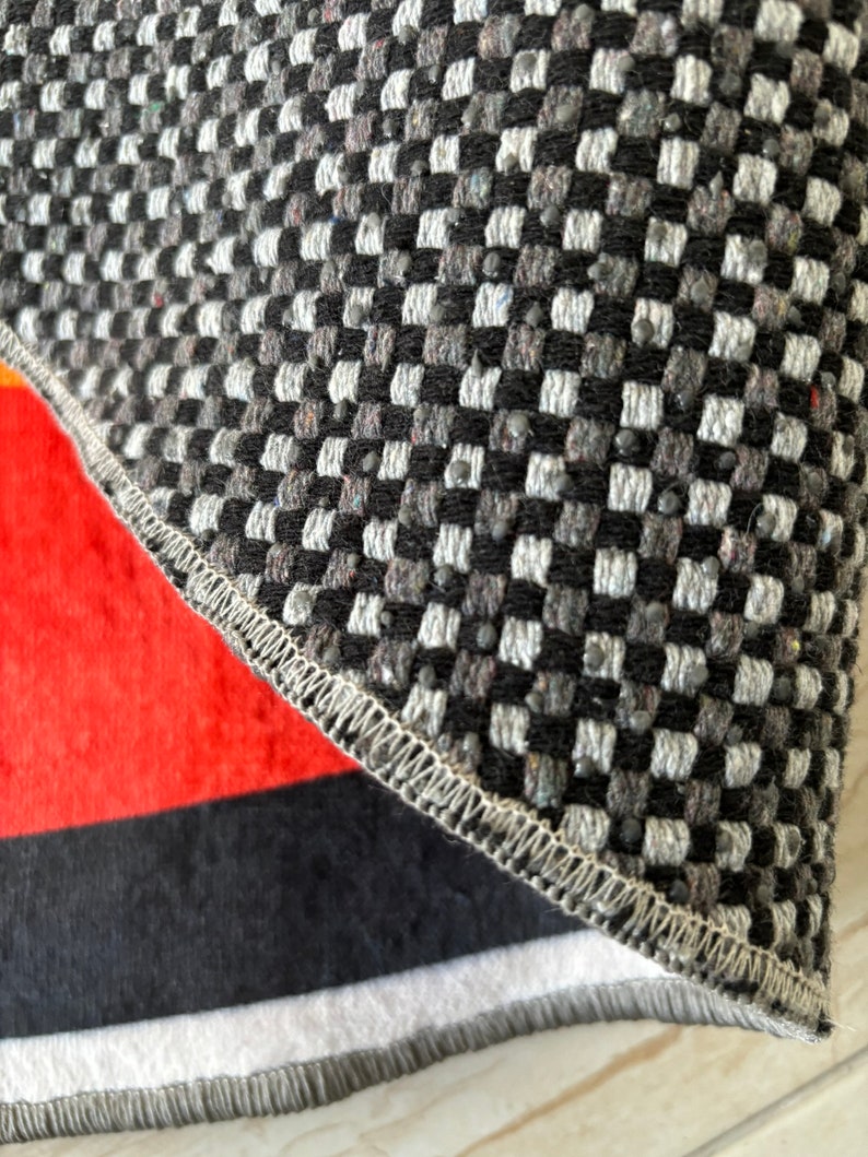 May include: Close-up of a section of a rug with a geometric pattern. The top portion features a black, white, and gray checkered design. The bottom section has a red, black, and white striped pattern. The edges are finished with a stitched border.