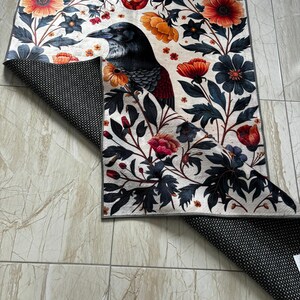 May include: A white rug with a black raven and colourful floral design. The rug has a black non-slip backing.