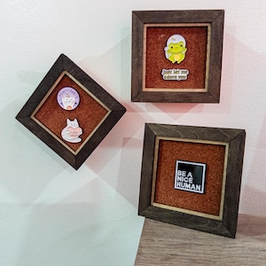 May include: Three framed pin displays on a white wall. Each frame has a dark wood finish and a corkboard backing. The pins include a cat with a heart, a frog in an egg, and a black and white square that says "BE A NICE HUMAN".