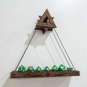 May include: A wooden dice shelf with a triangular design, suspended by chains. The shelf holds seven green dice with white numbering. The shelf is made of dark wood and is mounted on a white wall.
