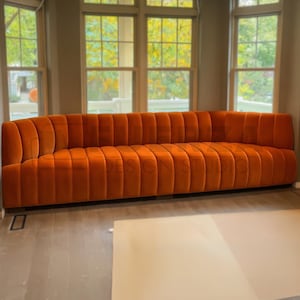 May include: An orange velvet sofa with a curved back and vertical channel tufting. The sofa is long and low, with a modern design. The room has large windows and a neutral color palette.