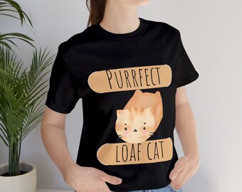 Op de afbeelding: Zwart T-shirt met een cartoon oranje kat die in een broodvorm is opgerold. De tekst "Purrfect Loaf Cat" is op het shirt gedrukt.
