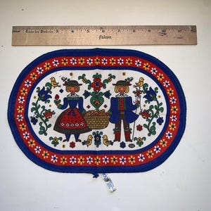 Può includere: Tovaglietta ovale in tessuto vintage con bordo blu e motivo floreale rosso e giallo. Il centro presenta un design di arte popolare di due figure in abiti tradizionali, circondate da fiori, uccelli e un cesto. La tovaglietta misura circa 28 cm di lunghezza.