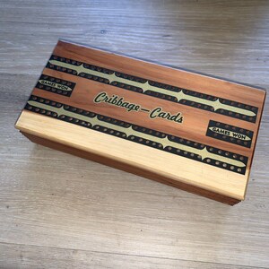 May include: A rectangular wooden cribbage board featuring a natural wood grain finish with two inlaid brass scoring tracks. The board displays the text "Cribbage-Cards" in the centre and "GAMES WON" on two separate black rectangular inlays.