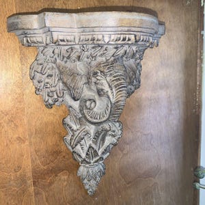May include: A decorative wall shelf with an elephant design. The shelf is made of a light-colored material with a distressed finish. The elephant is carved in relief, with intricate details. The shelf has a curved top and a decorative bottom.